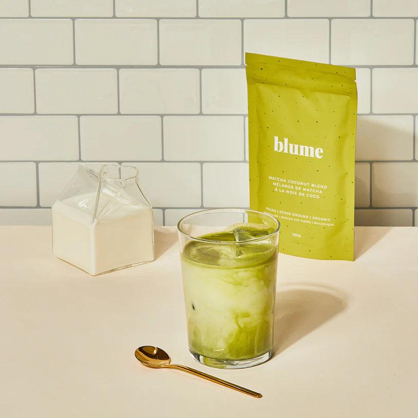 A glass of iced matcha latte with a spoon sits on a beige surface. A small glass bottle of milk is to the left. A green package labeled blume is propped up at the back. The background is a white tile wall.